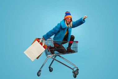 Side view of excited young female in bright clothes carrying paper bags like superhero while riding shopping cart against blue background clipart