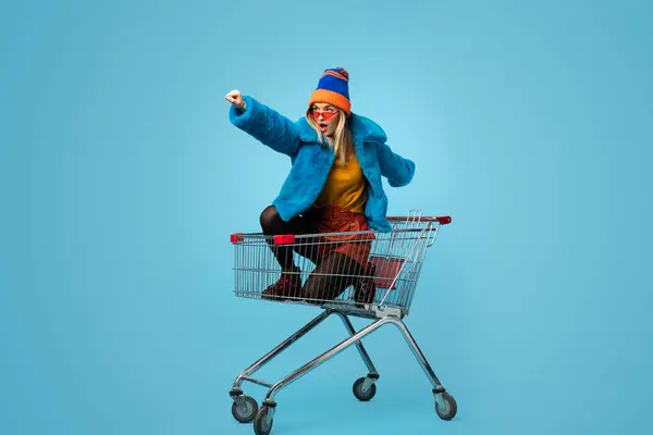 Side view of crazy trendy young female in bright clothes holding paper bags and riding shopping cart in superhero pose against blue background