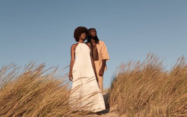 Saçları Afro saçlı kız arkadaşının yanında duran, bulutsuz mavi gökyüzüne karşı uzun beyaz elbiseler giyen ve gözlerini kaçıran Afrikalı genç bir adam.