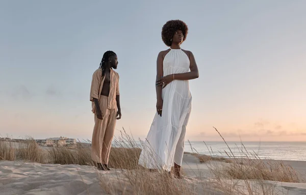身穿米黄色衣服的体形矮小的年轻非洲男子站在女友身后 身穿白衣 手挽手 日落时分站在海浪中的沙滩上 — 图库照片