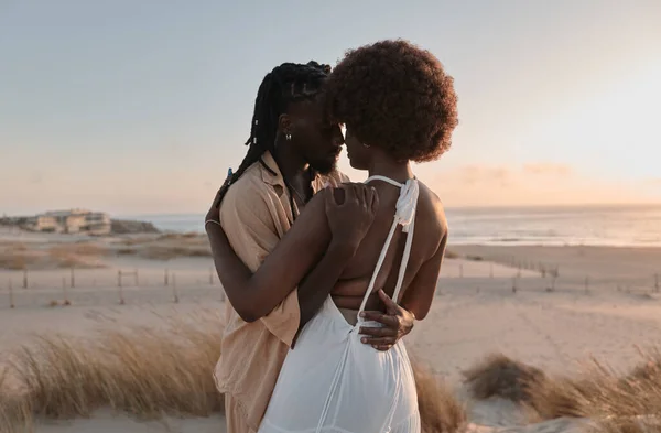 Young black man embracing girlfriend in white dress with Afro hair standing on sandy shore near waving sea against sundown sky during romantic trip