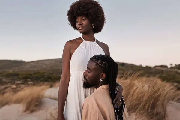 非洲年轻的性感孕妇 身穿白色连衣裙 抱着丈夫 跪在地上 抱着肚子靠着草山 — 图库照片