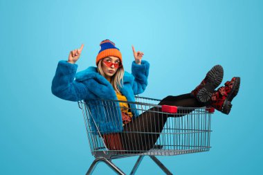 Mavi arka plan karşı alışveriş arabası otururken yukarı işaret renkli kıyafetli garip genç bayan