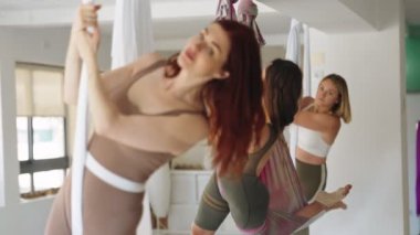 Aktif kıyafetli üç kadın, iyi aydınlatılmış bir stüdyoda, asılı hamakları kullanarak yerçekimi yoga egzersizleri yapıyorlar.