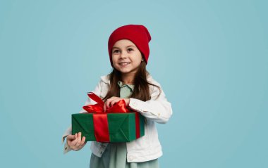 Moda kıyafetli mutlu kız, gülümsüyor ve mavi arka planda kırmızı kurdeleyle doğum günü hediyesi kutusunu açıyor.