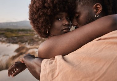 Afro saçlı ve dövmeli kız arkadaşını kucaklayan genç Afrikalı bir adam romantik bir tatil sırasında deniz kıyısında dağa karşı duruyor.