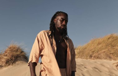 Gündüz vakti kumlu kumsalda kuru çimlerle bulutsuz mavi gökyüzüne karşı dururken, sıradan giyinmiş ve gözleri kapalı, kendinden emin genç Afro-Amerikan erkeklerinden.