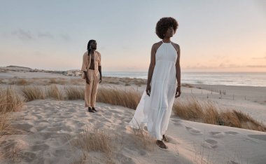 Gün batımında dalgalı denize karşı beyaz elbiseli bir kadının arkasında kumlu sahilde dikilen Afrikalı bir adam.