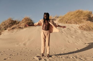 Genç, sakallı Afrikalı Amerikalı bir erkek vücudu, çıplak ayakla kumlu kumsalda ayakta dikilirken, güneşli bir günde kuru çimenler ve bulutsuz mavi gökyüzüne karşı gölgeli.