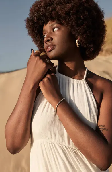 Young tender African female model with brown Afro hair standing on sandy shore against cloudless sky on sunny summer day