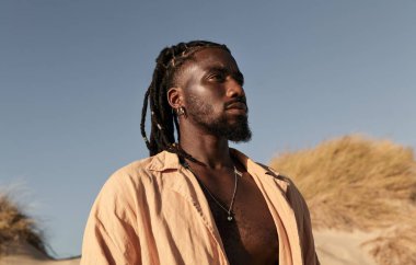 Ciddi, sakallı genç bir Afrikalı erkek yaz günü kuru çimlere ve bulutsuz mavi gökyüzüne karşı duruyor.