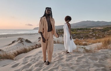 Tüm vücudu, kumlu sahil erkeğinin üzerinde çıplak ayakla dikilip bulutsuz gün batımına karşı gözlerini kaçıran genç Afrikalı Amerikalı çiftlerle dolu.