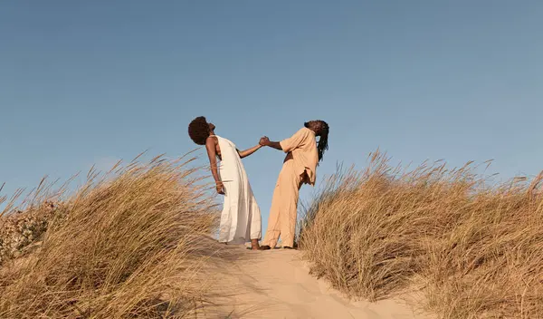 Full body of young African woman in white dress holding hands with boyfriend leaning back, while standing on sand near dry grass against cloudless blue sky