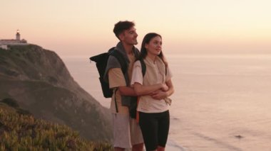 Bir çift genç gezgin, kadını arkadan kucaklayan bir adam, ikisi de sırt çantalı, çarpıcı bir uçurumun kenarında gün batımında rahatlatıcı bir anın tadını çıkarıyorlar..