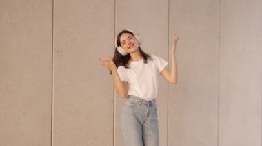 Kot pantolonlu ve beyaz tişörtlü genç bir kadın en sevdiği şarkıyla enerjik bir şekilde dans ediyor, kulaklık takıyor, arka planında beton bir duvar var.