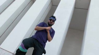 Sokak kıyafetleri ve güneş gözlüklü genç bir erkek, minimalist şehir geçmişine karşı dinamik hareketlerle çağdaş modern dans ediyor.