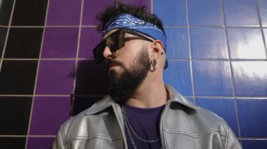Saç bandı takmış hip-hop sanatçısı ve güneş gözlüklü parlak duvar arkaplanındaki tavırlarıyla başını çeviriyor.