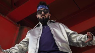 Parlak ceketli ve güneş gözlüklü rapçinin şehir sokaklarında rap el hareketlerini gösteren düşük açılı çekimi.