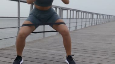 Spor kıyafetleri içinde görülmeyen bir kadın, yan ayak basamaklarında, açık havada tahta bir kordonda direnç bandıyla başarılı olur.