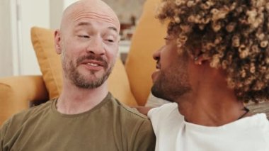 Yakın plan çekimde, kel beyaz bir erkek ve Afrikalı bir kıvırcık erkek, sevgi dolu bir eşcinsel çift, evlerinin rahatlığında sohbet eder ve gülümsemelerini paylaşırlar.