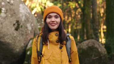 Sarı şapkalı ve yağmurluklu genç bir kadın, seyahat çantası taşıyor, gülümsüyor ve yemyeşil bir ormanı gezerken kameraya bakıyor.