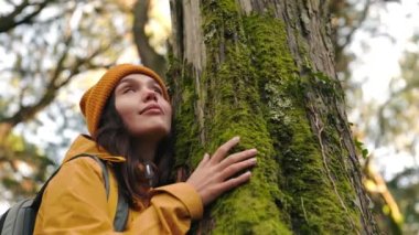 Güzel, genç bir kadın, kalın giysiler içinde, sırt çantasıyla, bir ağaca sarılıyor ve dinliyor, bereketli bir ormanda, doğanın dingin güzelliğine dalmış.