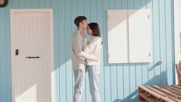 晴れた日の明るい青いビーチハウスに立っている若いカップルの柔らかい瞬間 キスを共有し 新しい家に移ることを象徴する愛を表現します — ストック動画