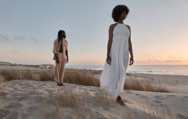 Beyaz elbiseli kız arkadaşının arkasında kumlu tepede gün batımında gökyüzünün altında denize karşı duran Afrikalı genç bir adam.