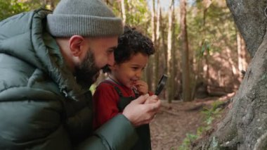 Ceketli ve şapkalı bir babanın, şirin, kıvırcık oğluyla birlikte büyüteç kullanarak ormanı gezdiği sıcak bir sahne.