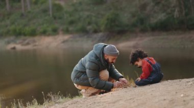 Bir baba ve oğulun göl kenarında neşeli bir şekilde kumları bir sopa ve büyüteçle keşfettikleri karıncaları ve doğanın harikalarını keşfettikleri iç açıcı bir sahne.
