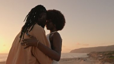 Aşk ve tutku kavramı, beyaz elbiseli çarpıcı siyahi bir kadın güneş batarken kumsalda rastalı yakışıklı bir Afrikalı erkeği kucaklıyor ve yoğun bir bağ kuruyor.