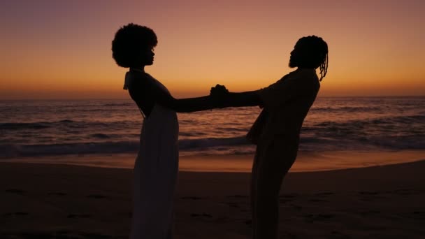 在夕阳西下的温暖光芒中 一个年轻的非洲女人和一个英俊的黑人男人在海浪的背景下手牵手 表达了他们的信任和联系 — 图库视频影像