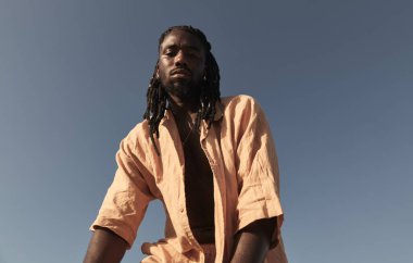 Güneşli bir havada mavi bulutsuz gökyüzüne bakan ve kameraya bakan uzun saçlı Afrikalı genç adamın alçak açısı.