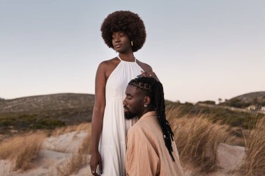 Diz çöküp beyaz elbiseli kız arkadaşına sarılan genç, şehvetli Afrikalı adam gün batımına karşı kuru otlak tepelerde duruyor.