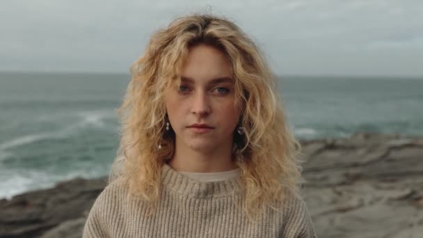 風の強い日に岩場の海岸で深く熟考したカメラを眺めながら セーターに乗った若い金髪の女性の魅惑的な肖像画 — ストック動画