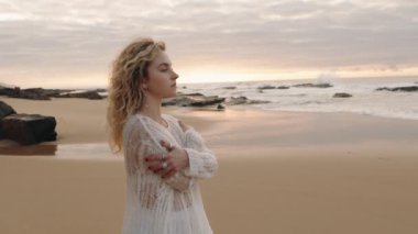 Beyazlı genç bir kadın sahilde tek başına duruyor, kendine sarılıyor ve gün batımında okyanusu seyrediyor.