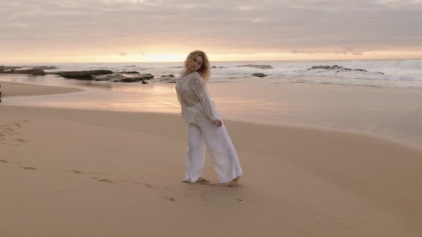 白い服の美しい若いブロンドの女性がビーチでゆっくりとした動きで踊り 衝突波の魅惑的な背景に囲まれています — ストック動画