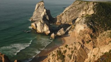 Güneş batarken Portekiz 'deki Praia da Ursa' nın nefes kesici güzelliği baş döndürücü kayalıklara sıcak bir ışıltı saçıyor ve okyanus dalgalarının plaja çarpan ritmik dansı.