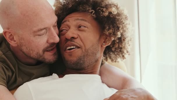在自然光下 一个英俊的白人男人从后面抱着一个黑发卷曲的男人坐在沙发上 庆祝同性爱情和团结 — 图库视频影像