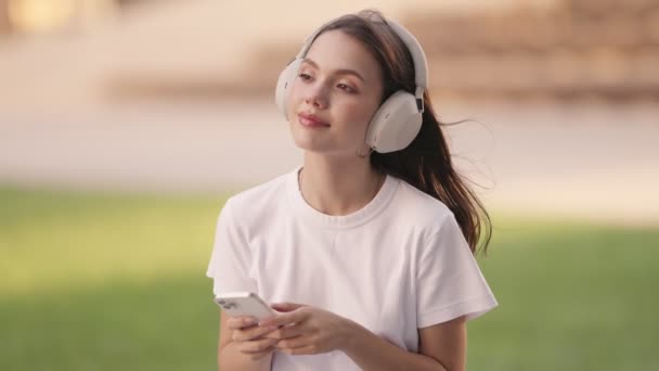 夏日的微风中 年轻女子沉醉在音乐中 戴着耳机 在智能手机上滚动播放音乐 — 图库视频影像