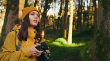 Sıcak ceketli ve sarı şapkalı genç bir kadın ormanı geziyor, profesyonel kamerasıyla anları yakalıyor.