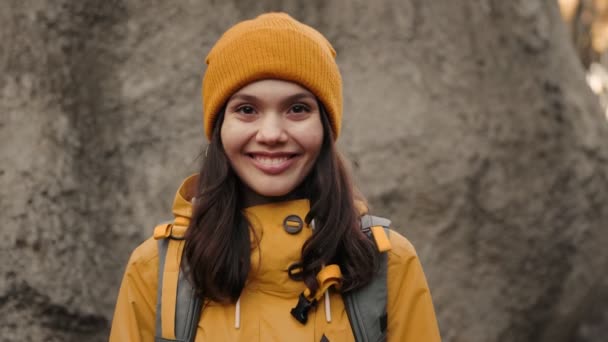 一位年轻的女徒步旅行者穿着温暖的黄色衣服 背着背包在森林里的一块巨石上对着摄像机微笑 — 图库视频影像