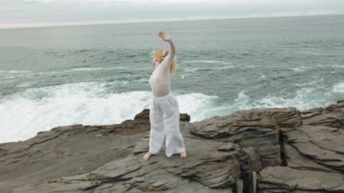 Beyazlı genç bir kadının zarafetine tanık olun. Kayalık bir uçurumun kenarında dans ediyor. Okyanus dalgalarıyla uyumlu.