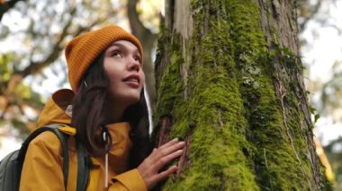 Yağmurluk giymiş genç bir kadın, ormanı keşfediyor ve yosun kaplı bir ağaca dokunuyor, büyüleyici çevreyle derin bir bağ hissediyor, ağaç gövdesini kucaklıyor.