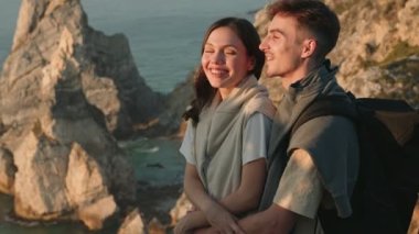 Genç bir adam güzel bir kadını kucaklar, kayalık bir uçurum üzerindeki büyüleyici okyanus manzarasının tadını çıkarır.