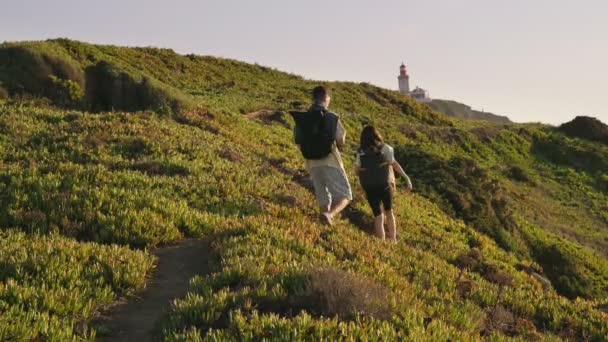 若いカップルが一緒にバックパックでハイキングトレイルを歩いて 魅力的な灯台につながります — ストック動画