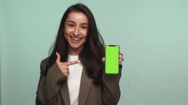 Akıllı ceketli neşeli genç bir kadın yeşil ekranlı cep telefonunu işaret ediyor. Tarafsız bir arkaplanda içeriğiniz için hazır..