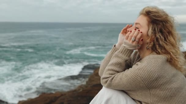 海のそばに座り 岩場にぶつかる波を見つめ 自然の中で孤独と反射を体現する高価なブロンドの女性 — ストック動画