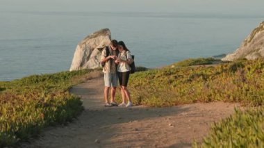 Genç bir çift, gün batımında sırt çantalarıyla sahil şeridini keşfederken akıllı bir telefonla gezinti macerasından zevk alır..