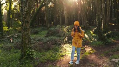 Sarı ceketli genç bir kadının büyüleyici görüntüsü yalnız başına yürüyüş yapıyor, yeşil bir ormanın dingin güzelliği içinde güneşleniyor..